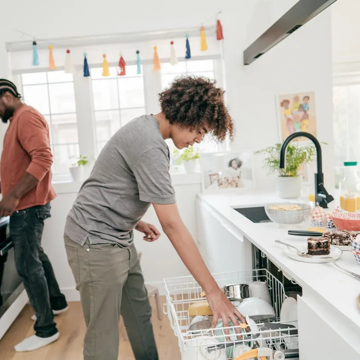 A teen loading a dishwasher in a bright kitchen with a parent in the background.