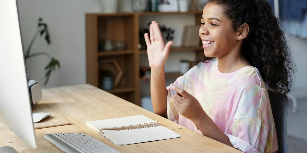 Young girl waving at a computer screen in a home office setting